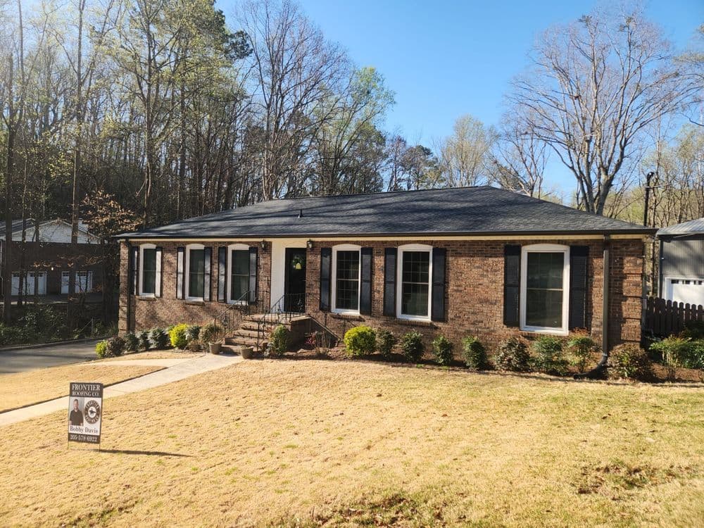Brick ranch-style home with lush yard and for sale sign, surrounded by trees.