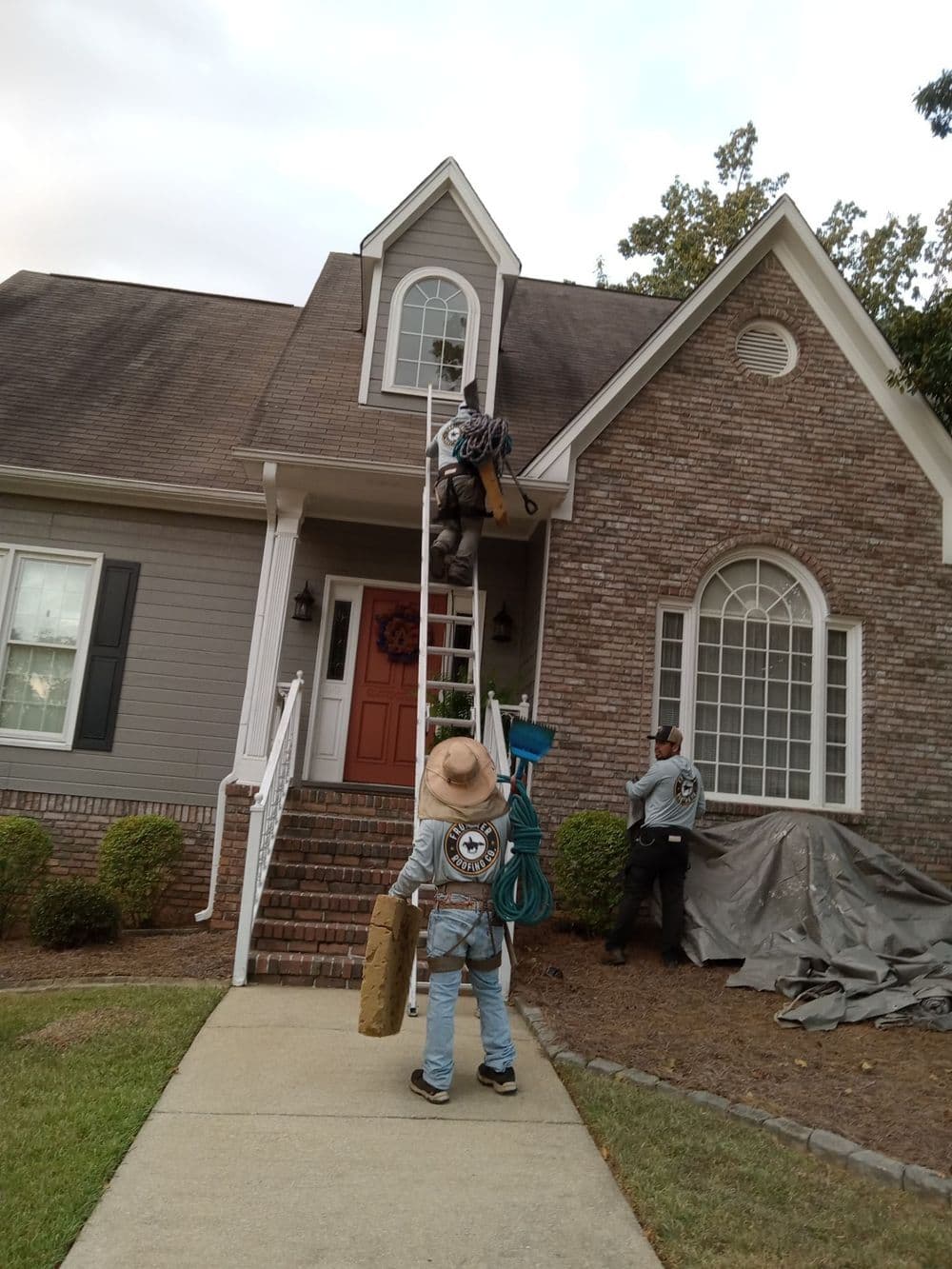 Workers using a ladder for roof repairs on a residential home with landscaping.