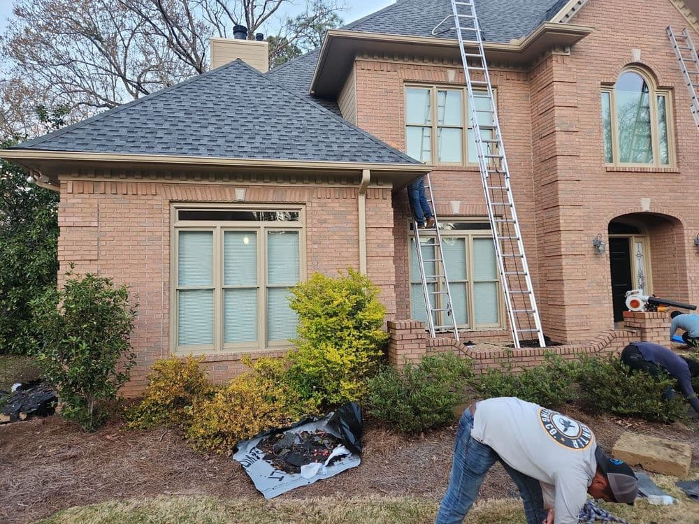 Workers performing roof maintenance on a brick house with ladders and landscaping.