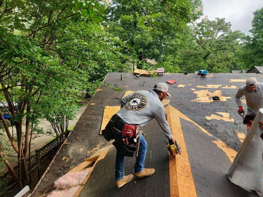 Roofer repairing a residential roof, with trees in the background and debris on the surface.