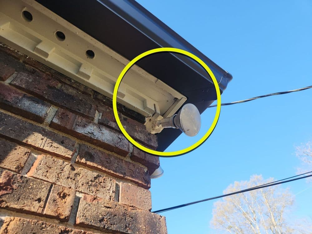 Security camera mounted on a brick wall under a roof eave, with blue sky in the background.