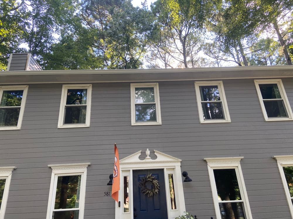 Gray two-story house surrounded by trees, featuring a front door wreath and an orange flag.
