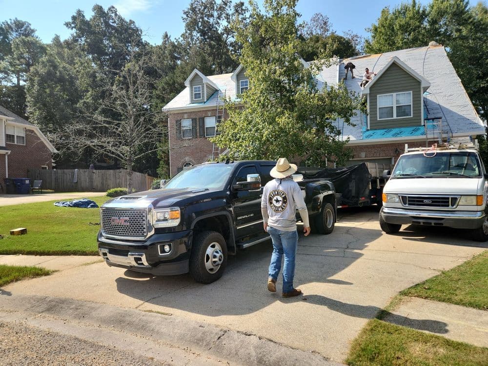 Man in cowboy hat overseeing roofing work on a house, with trucks parked nearby.