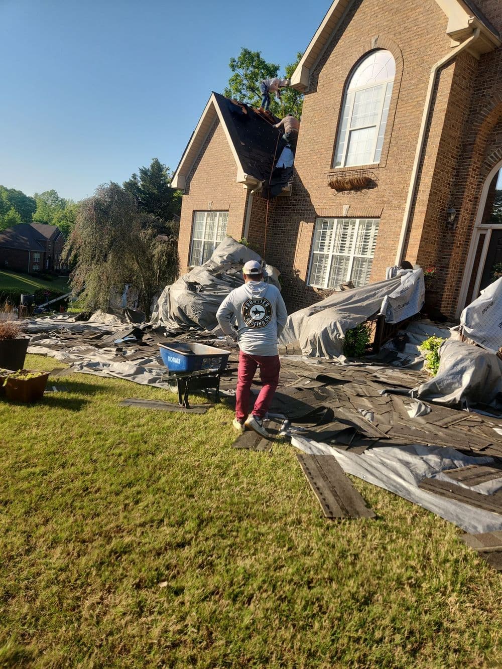 Roof repair work in progress on a brick house with tarps, worker supervising from the yard.
