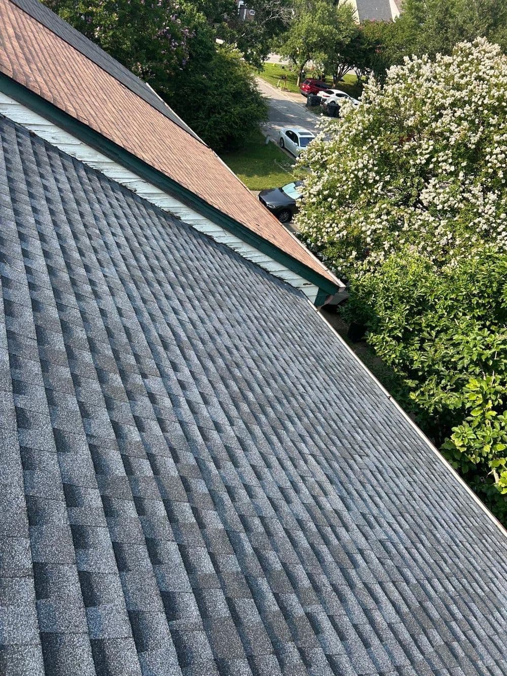Aerial view of a textured gray shingle roof with surrounding greenery and a car in the background.