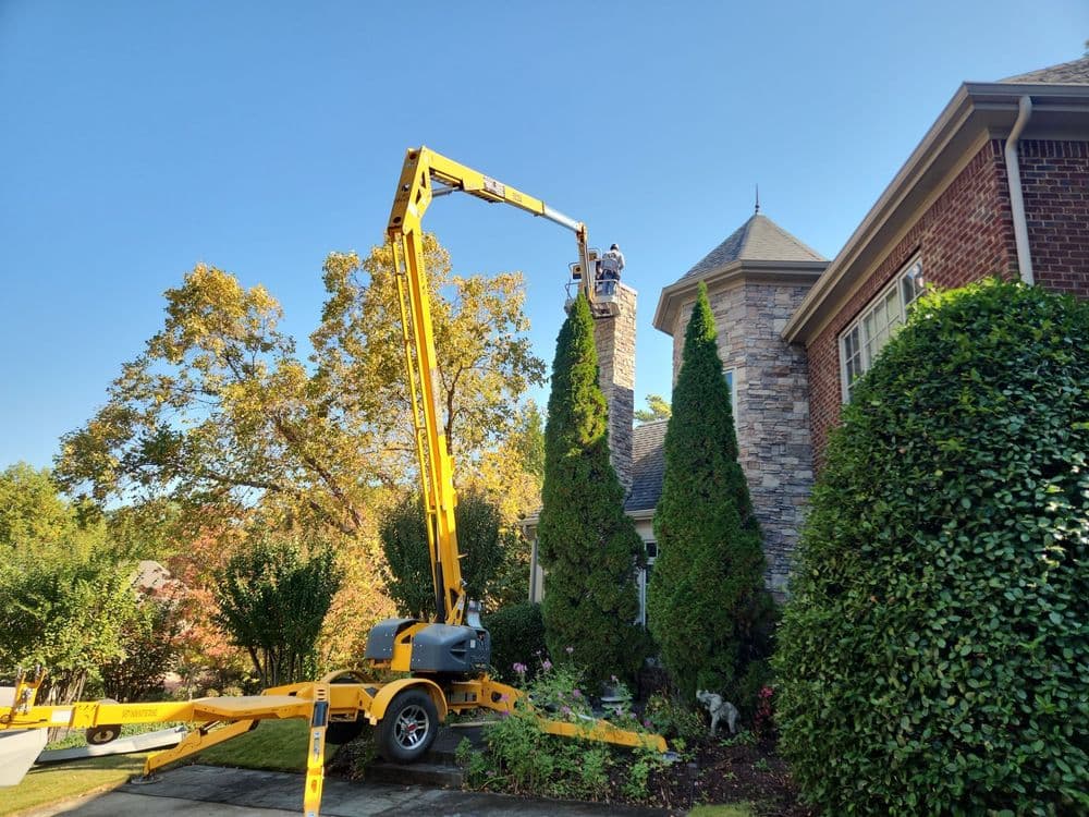 A tree service worker uses a lift to trim tall shrubs by a brick house under clear blue skies.