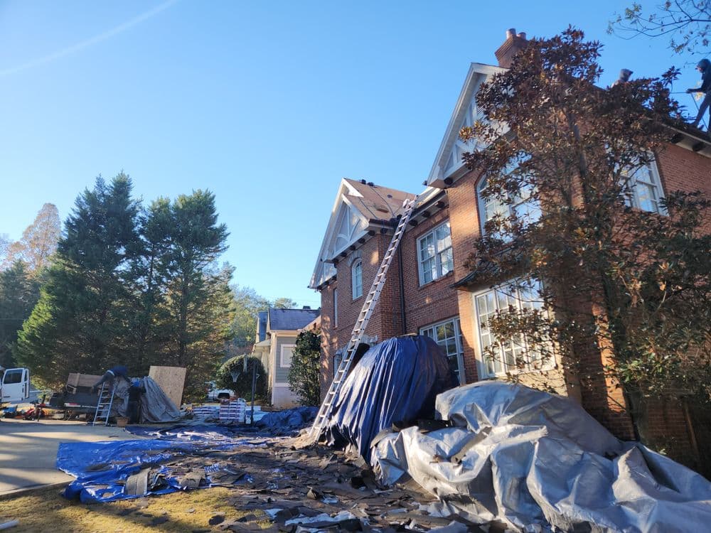 Roof repair in progress on a brick house, with ladders and tarps covering the ground.