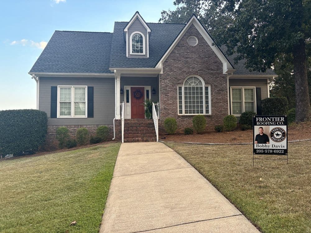Charming grey house with brick accents, lush landscaping, and a sign for Frontier Roofing Co.