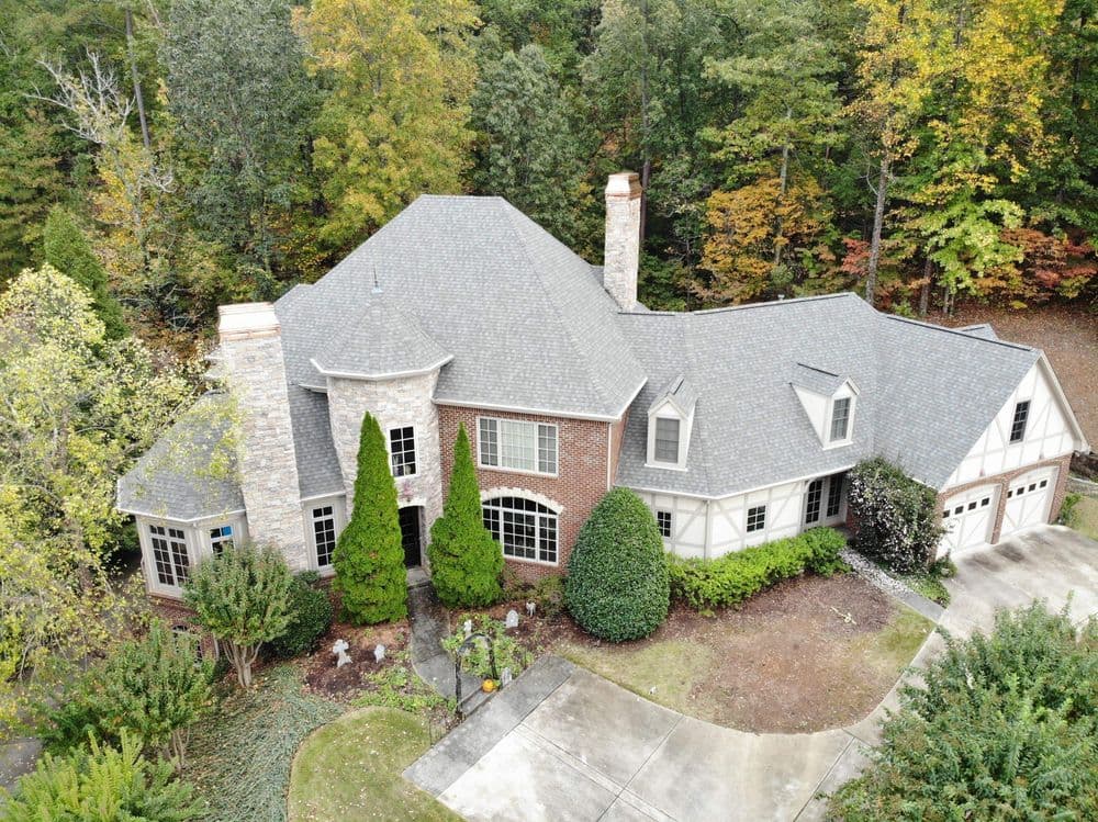 Aerial view of a large brick home with gray shingles, surrounded by green trees and landscaping.