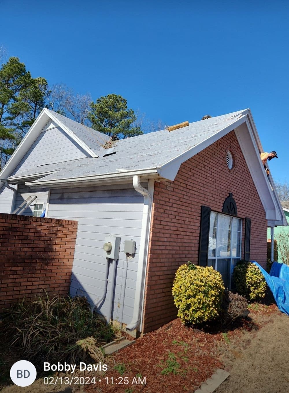 Roof repair in progress on a brick house, with a worker on the roof and sunny sky.