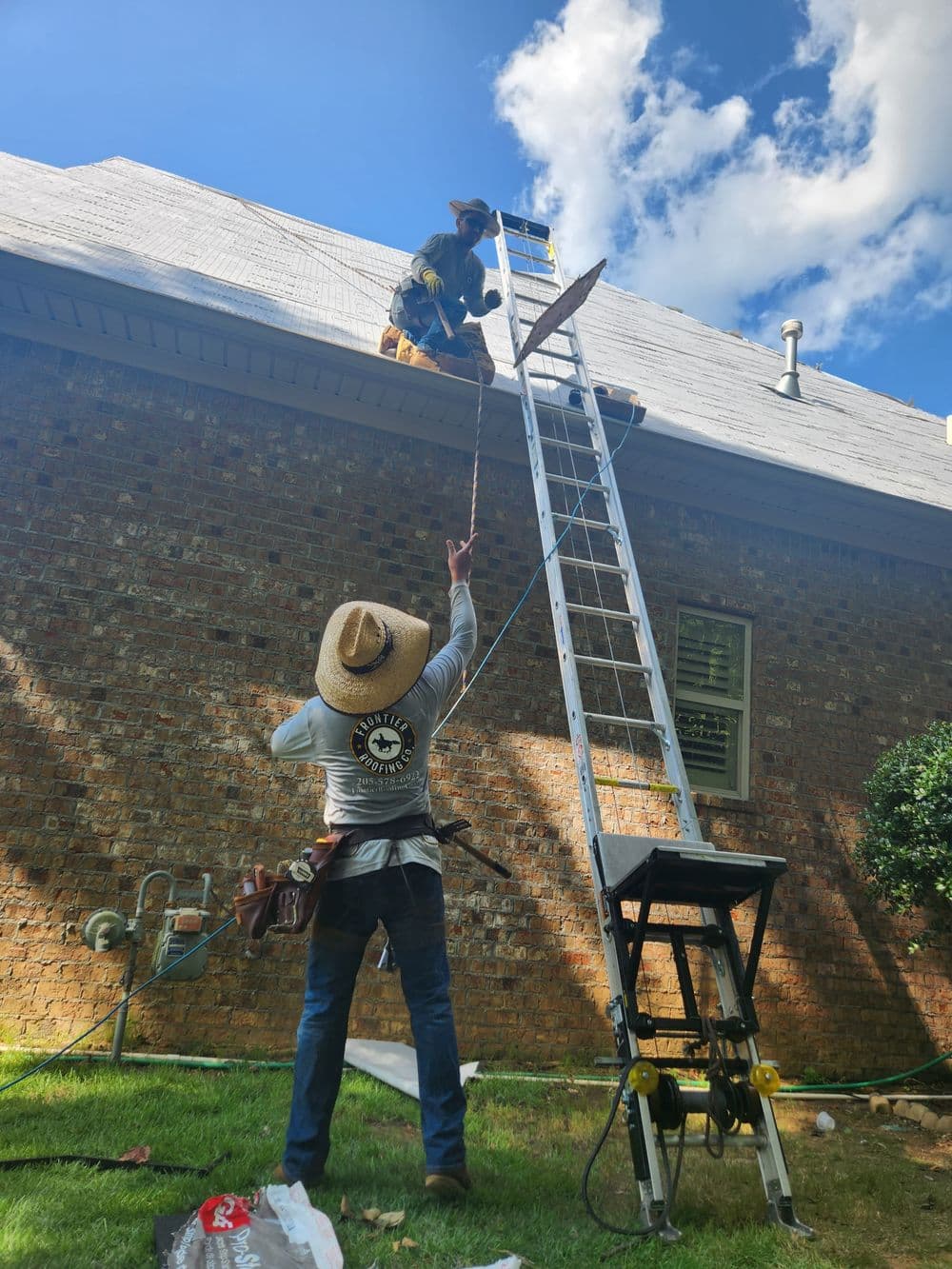 Workers repairing a roof using a ladder, with tools and materials on the ground.