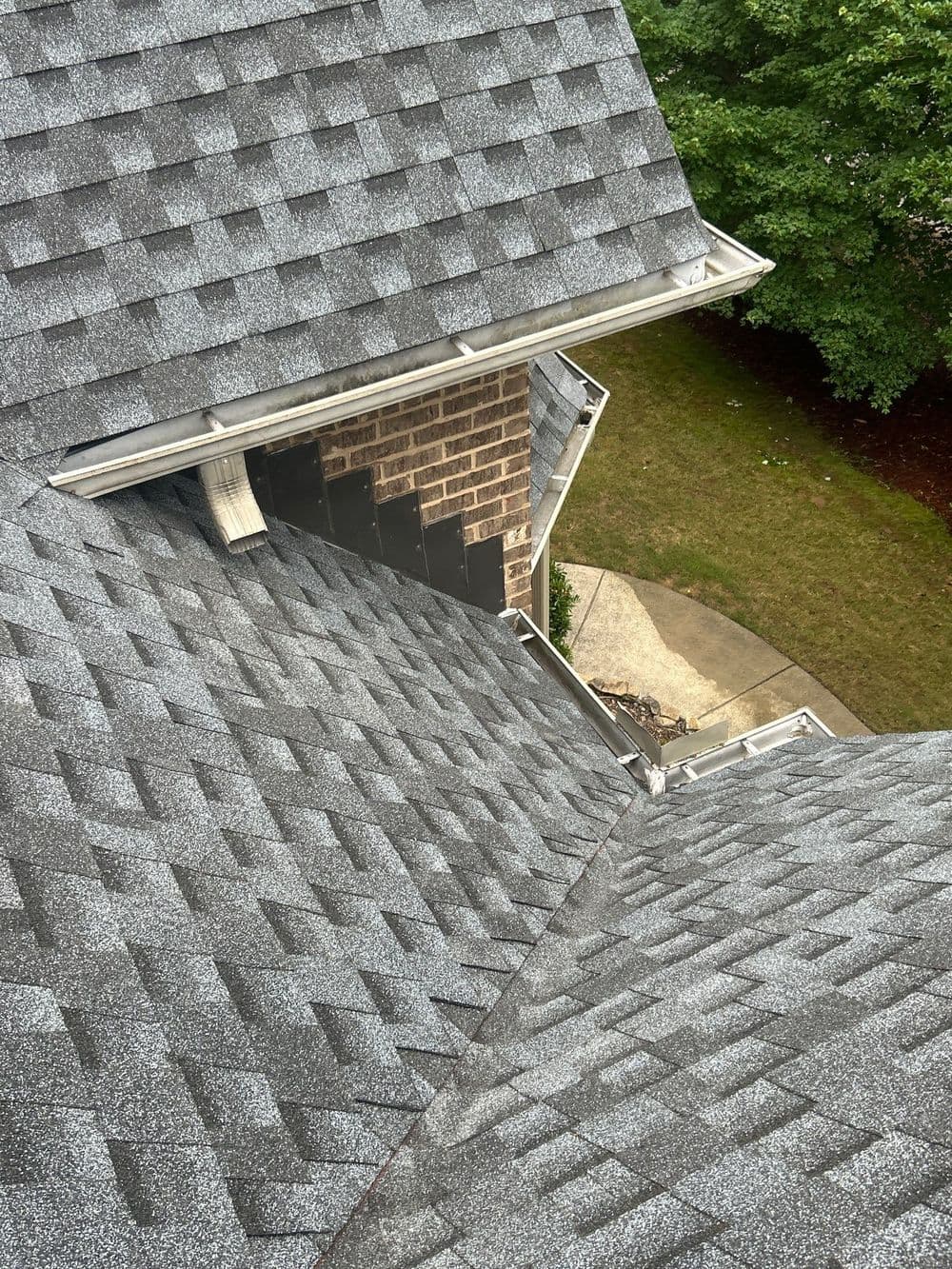 Aerial view of a gray shingle roof with a brick wall and green lawn in the background.