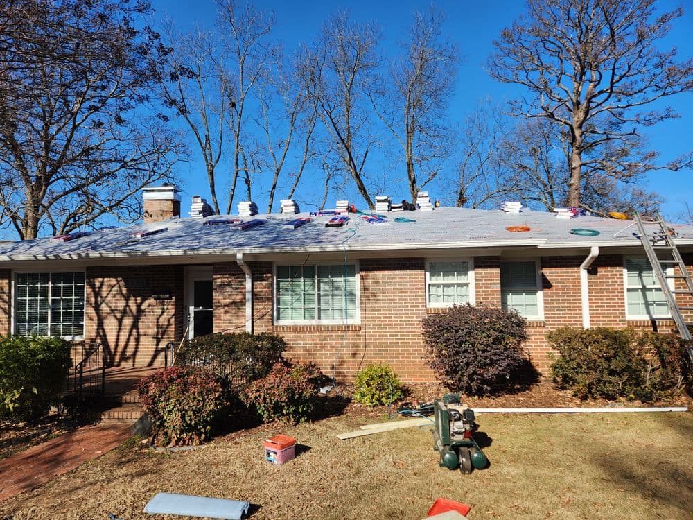 Roofing project in progress on a brick house surrounded by trees and tools. Clear blue sky.