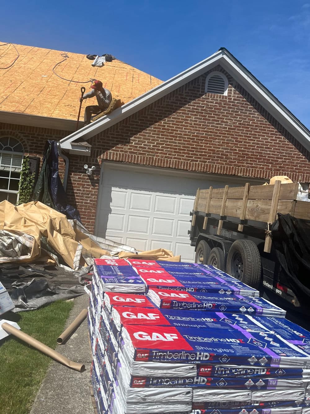 Roofing contractor installing shingles on a house with stacked GAF Timberline HD materials nearby.