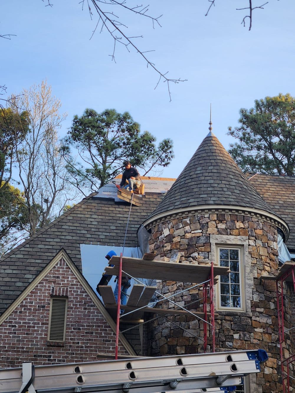 Roofers installing shingles on a stone house with scaffolding and trees in the background.