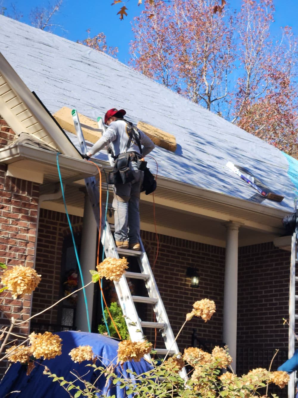 Roofer on ladder installing new roofing shingles on residential home with tools and materials.