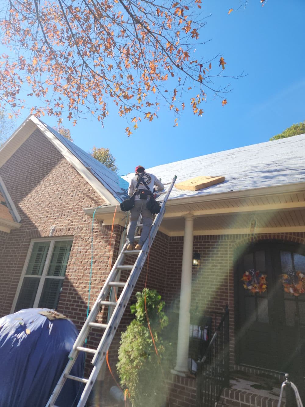 Worker repairing a roof with a ladder, autumn leaves on trees, and a brick house background.