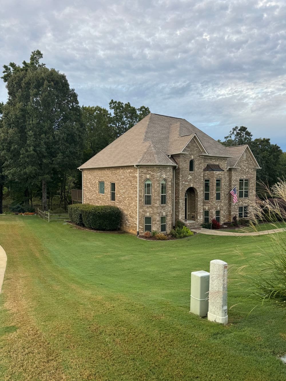 Elegant brick house with a peaked roof, lush green lawn, and American flag in a serene landscape.