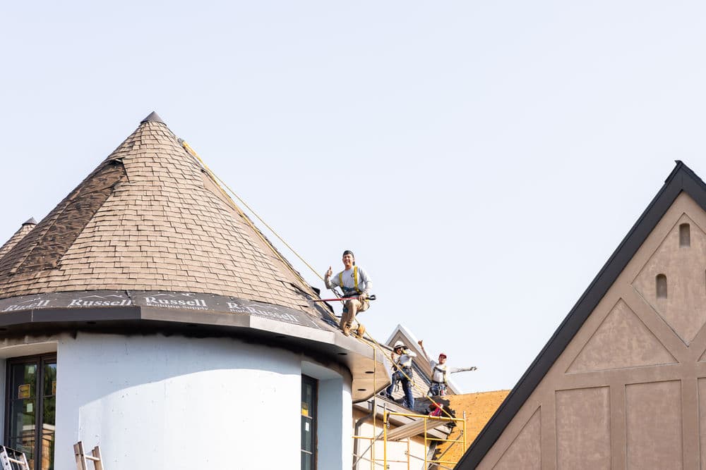 Construction workers installing shingles on a roof of a building with a turret design.