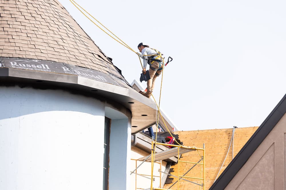 Roofing contractor working on a tall building with scaffolding, wearing safety harness.