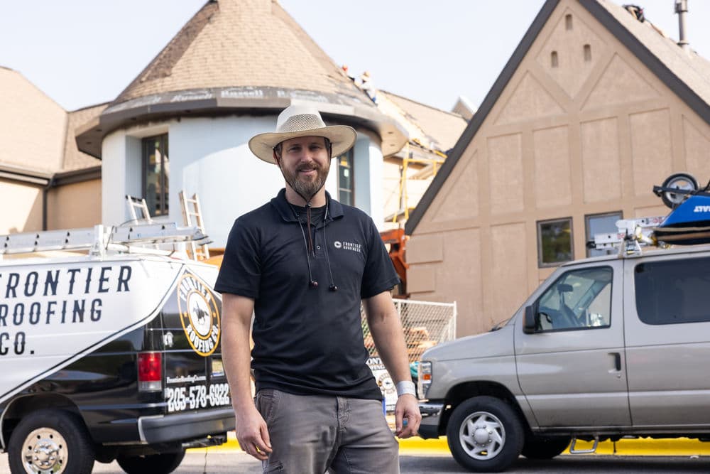 Roofer in a hat standing in front of a house undergoing roofing repairs with company vehicles.