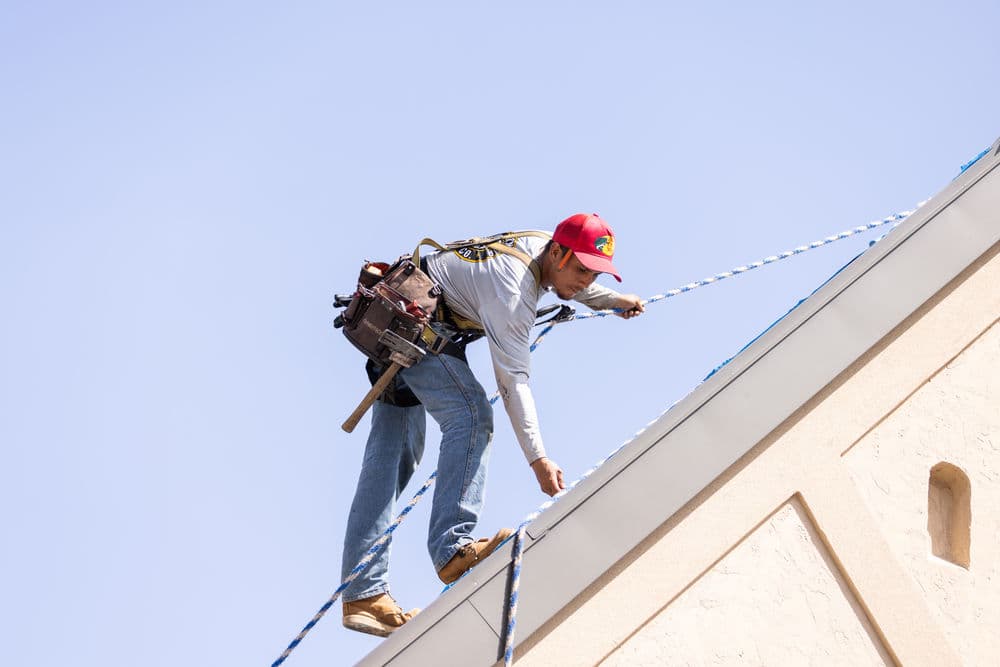 Roofer installing materials on a sloped roof against a clear blue sky. Safety harness visible.