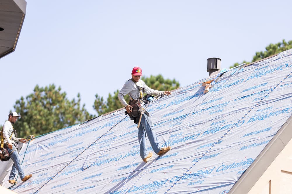Roofers installing protective sheeting on a residential roof under clear blue skies.