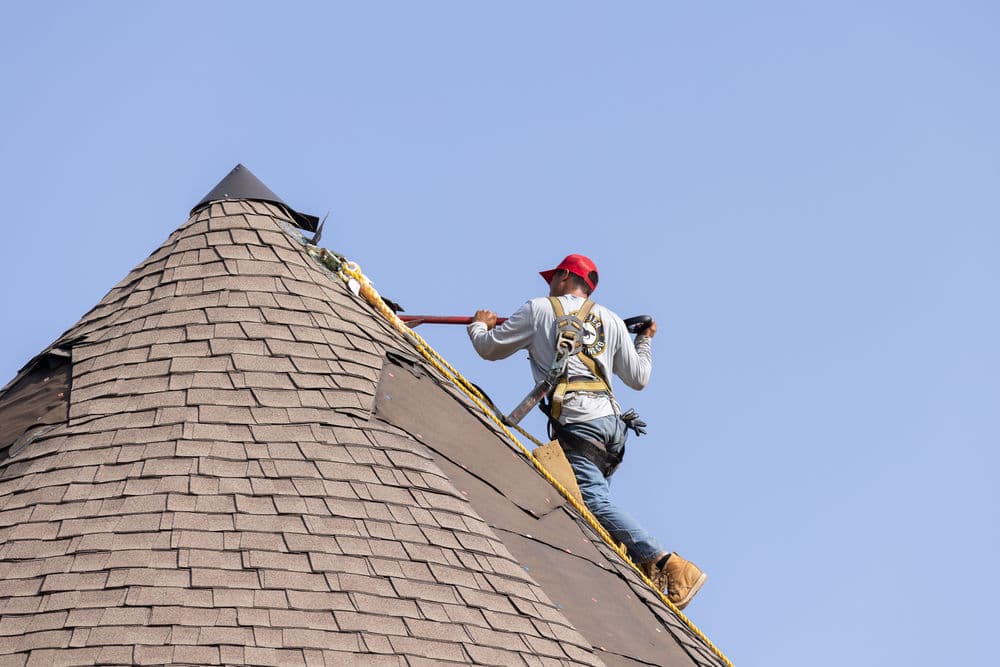 Roofer installing shingles on a steep, conical roof under clear blue sky.