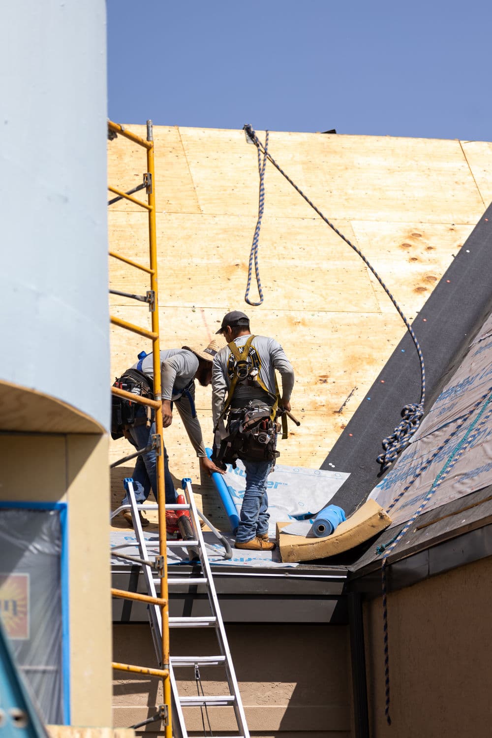 Construction workers installing roofing materials on a building under a clear blue sky.