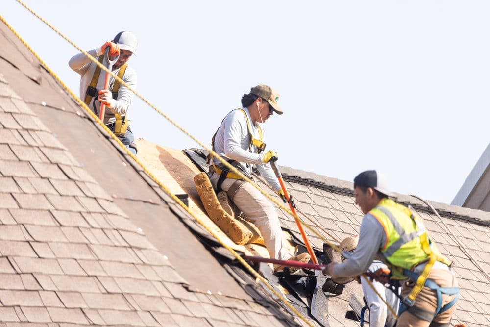 Construction workers repairing a roof with safety gear and tools on a sunny day.
