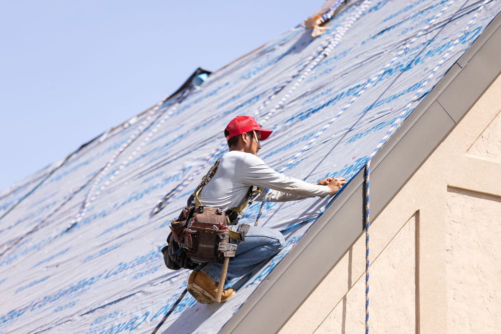Roofing contractor installing underlayment on a steep roof under clear blue skies.