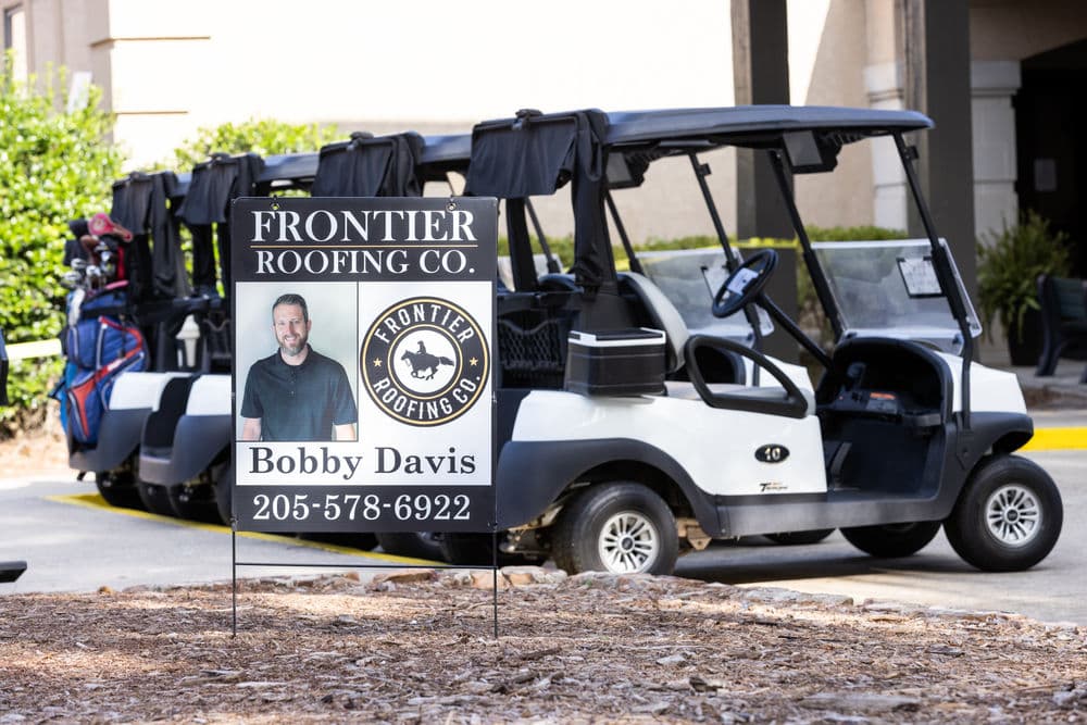 Frontier Roofing Co. sign featuring Bobby Davis near parked golf carts at an event.