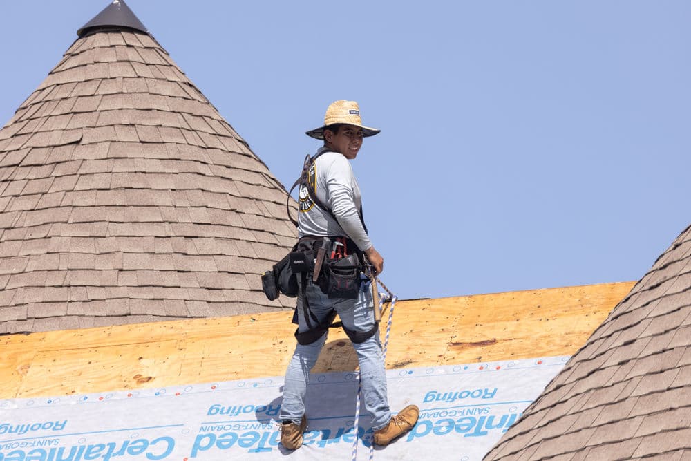 Roofing contractor installing shingles on a steep roof with a protective hat and tool belt.
